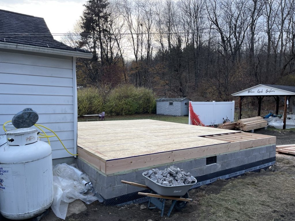 A construction site with trees in the distance. A small wheelbarrow full of rock debris sits next to a new foundation that is topped with a new bare wood floor. No walls or roof yet.
