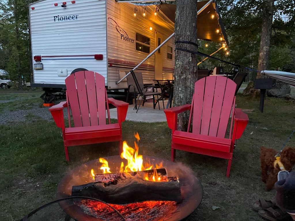 Two red Adirondack chairs face a campfire. Behind them sits a small travel trailer camper.